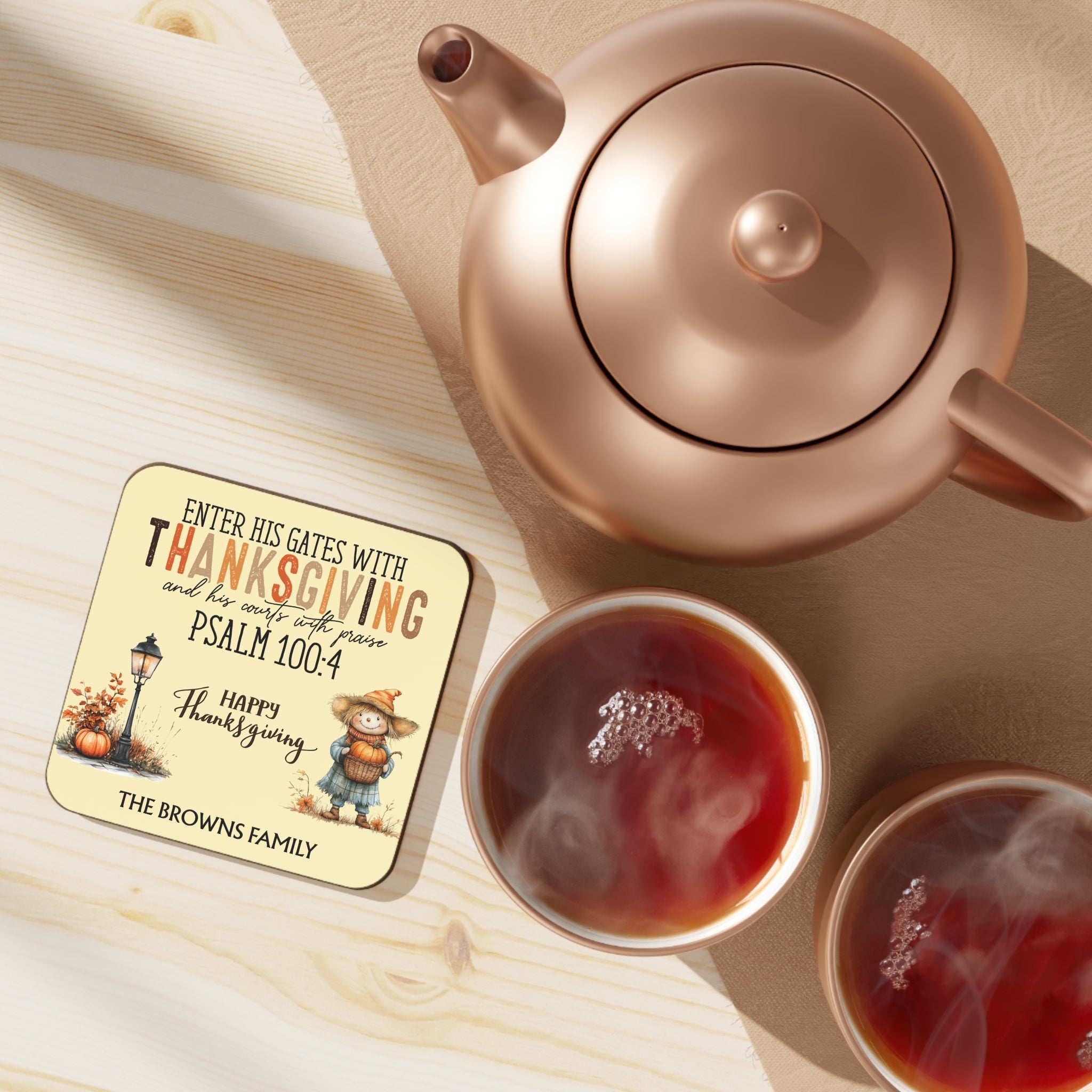 Rose gold teapot with two cups of tea on a wooden surface, featuring a Thanksgiving coaster.