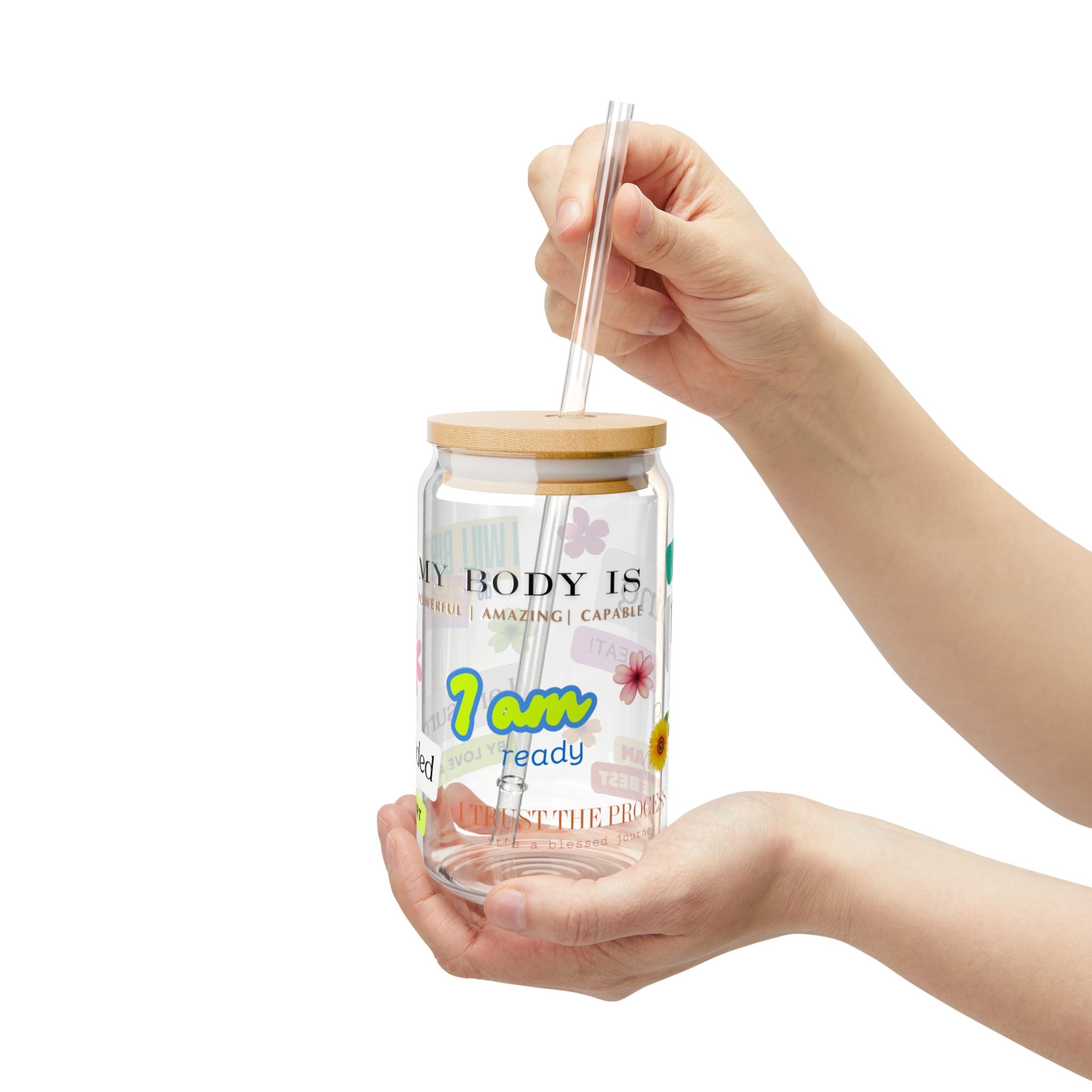 Hand holding a clear glass tumbler with a wooden lid and straw, featuring colorful text and graphics.