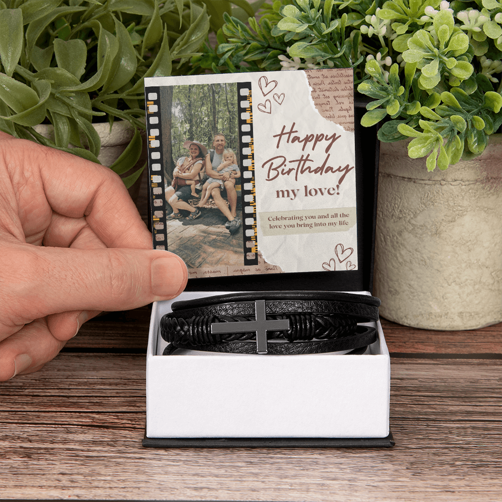 A black cross bracelet presented on a wooden box with a custom photo and message, displayed on a table with potted plants in the background.