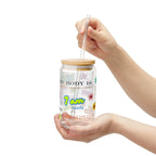 Hand holding a clear glass tumbler with a wooden lid and straw, featuring colorful text and graphics.