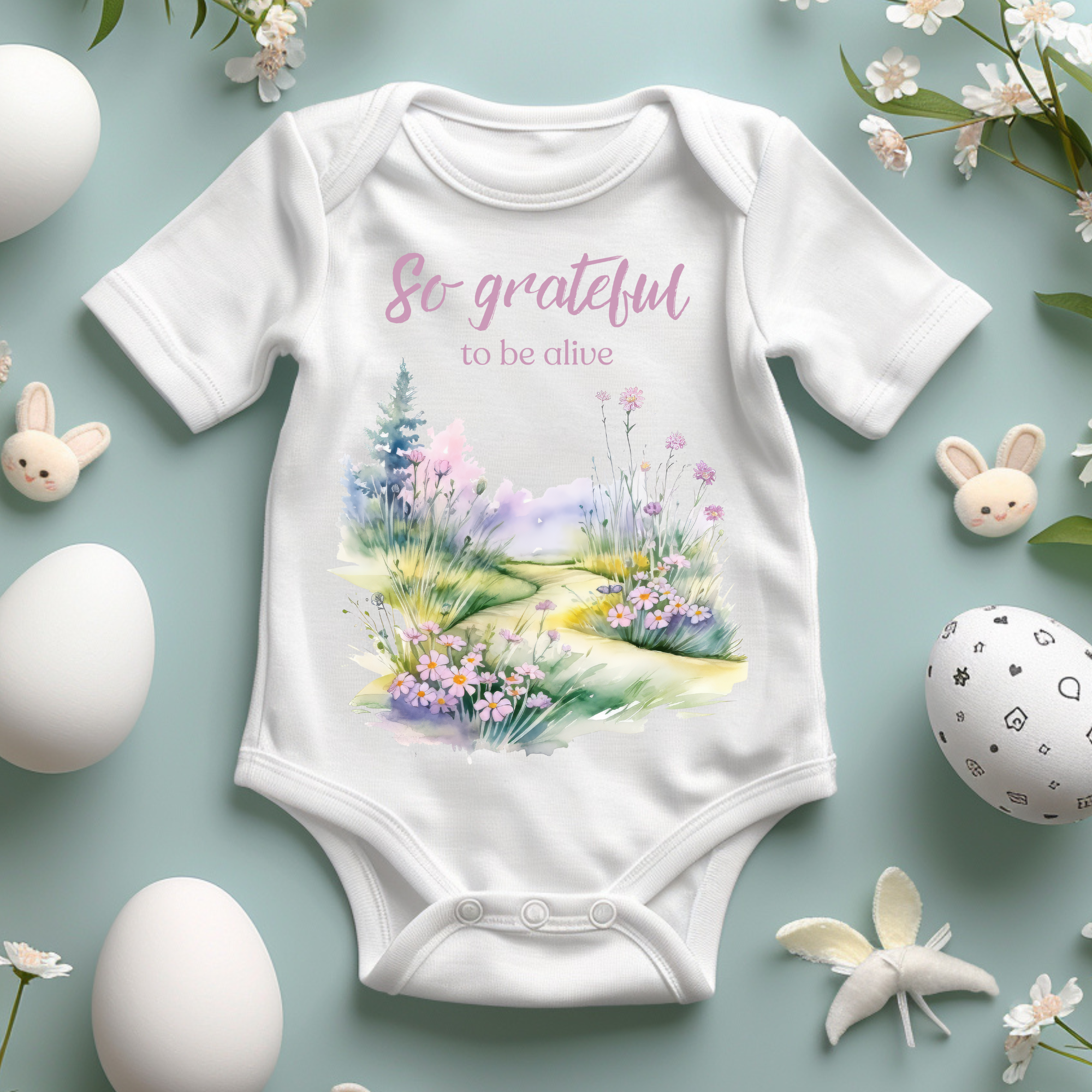 Baby onesie with "So grateful to be alive!" text and pastel nature design, made from soft 100% Airlume cotton, shown flat with envelope neckline and snap closures.