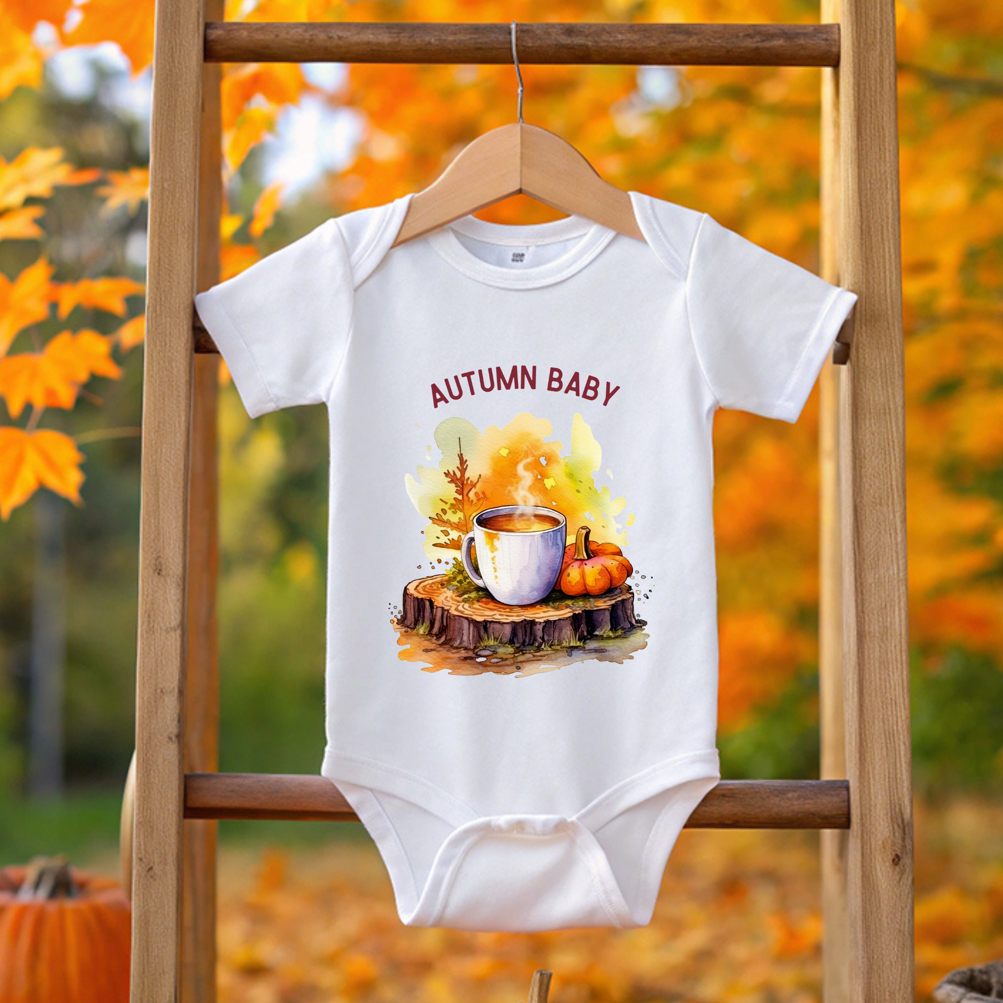 White baby onesie with 'Autumn Baby' design on a wooden ladder against an autumn background.