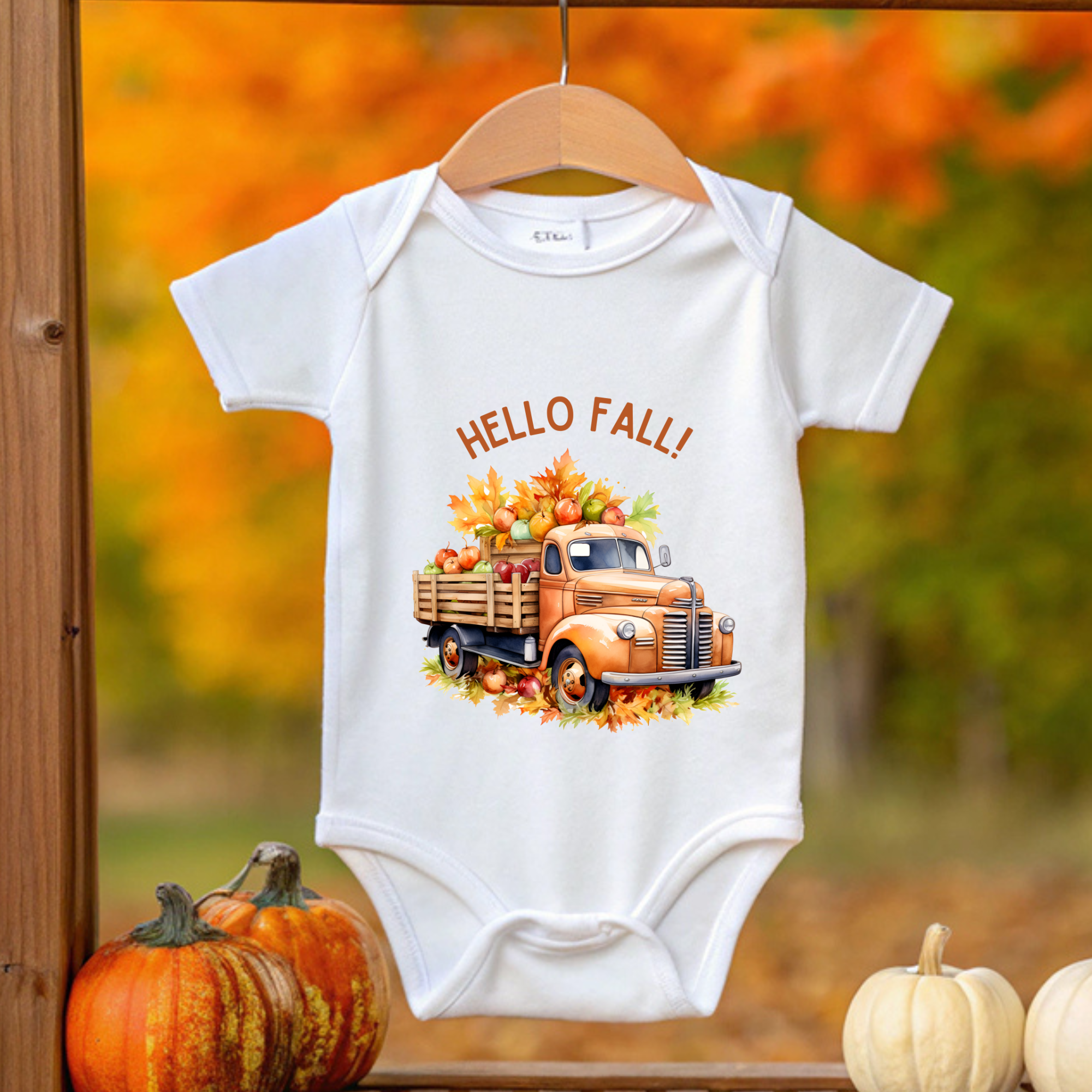 Baby onesie with a truck and 'Hello Fall!' text on a hanger against an autumn background.