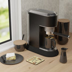 Black espresso machine on a wooden surface with a plate of cake and a window in the background.