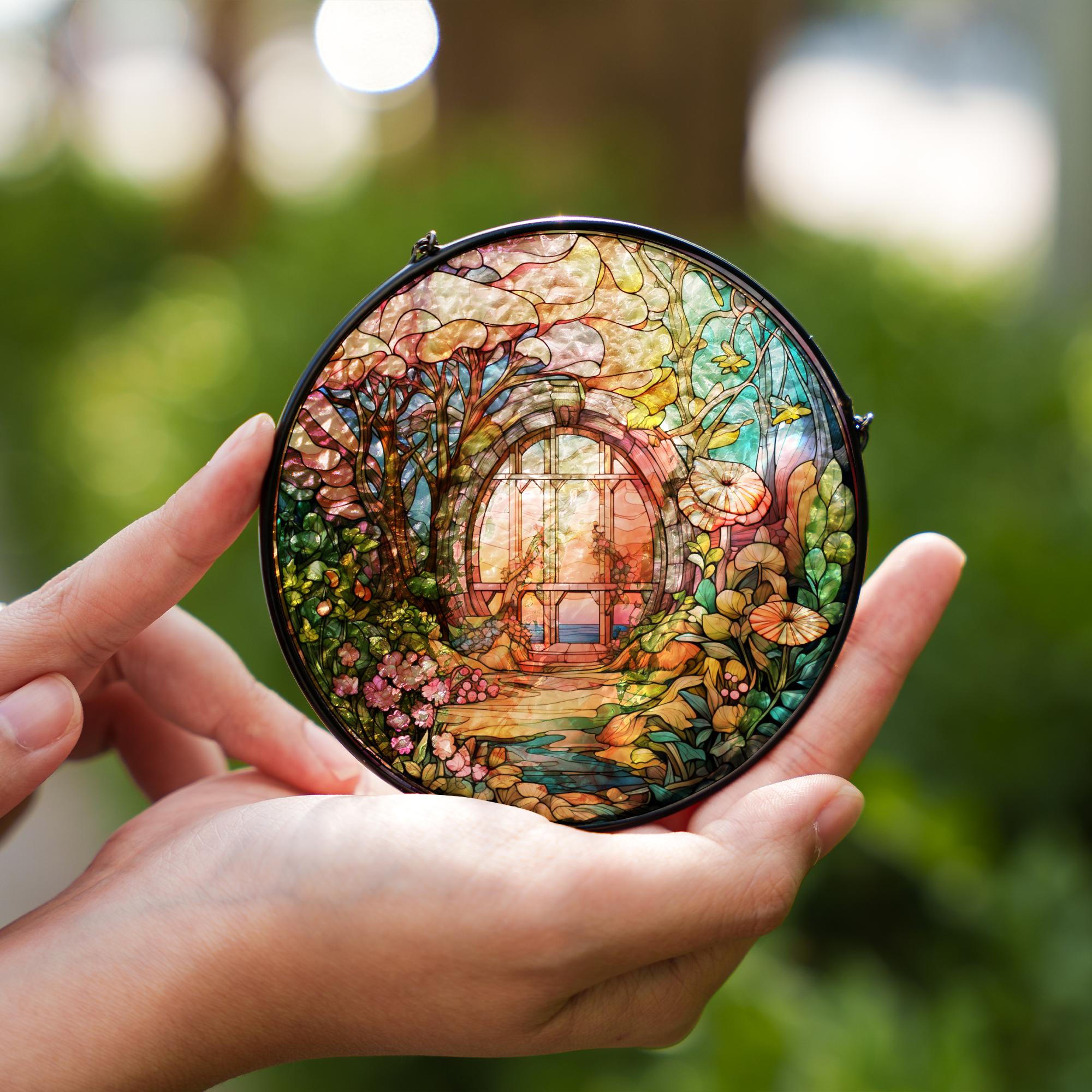 Hand holding a round, colorful stained glass suncatcher with a garden scene against a blurred green background.