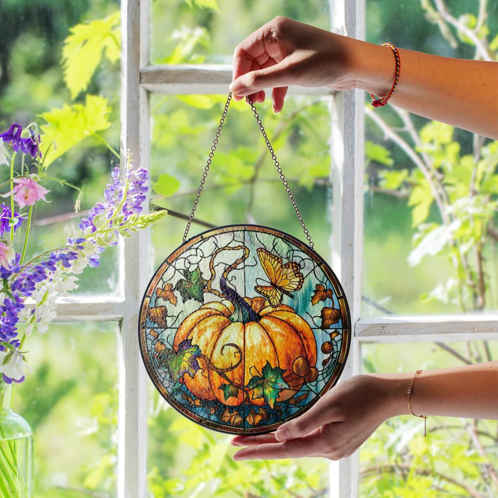 Stained glass pumpkin decoration held by a hand in front of a window with greenery.
