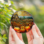 Round decorative item with a stained glass-style design of pumpkins and a house held by hands against a blurred green background.