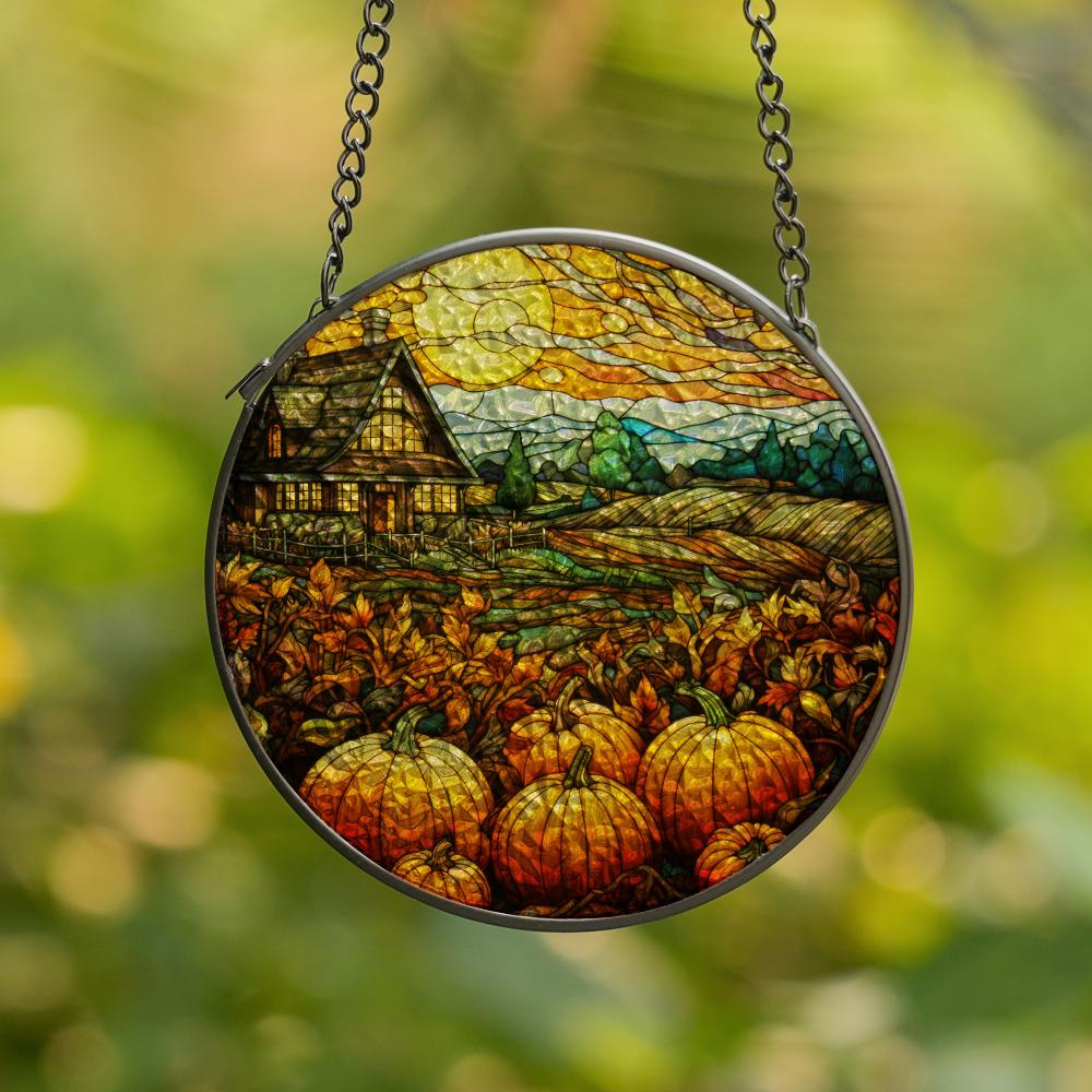 Round stained glass panel with autumn scene featuring pumpkins and a house, hanging against a blurred green background.