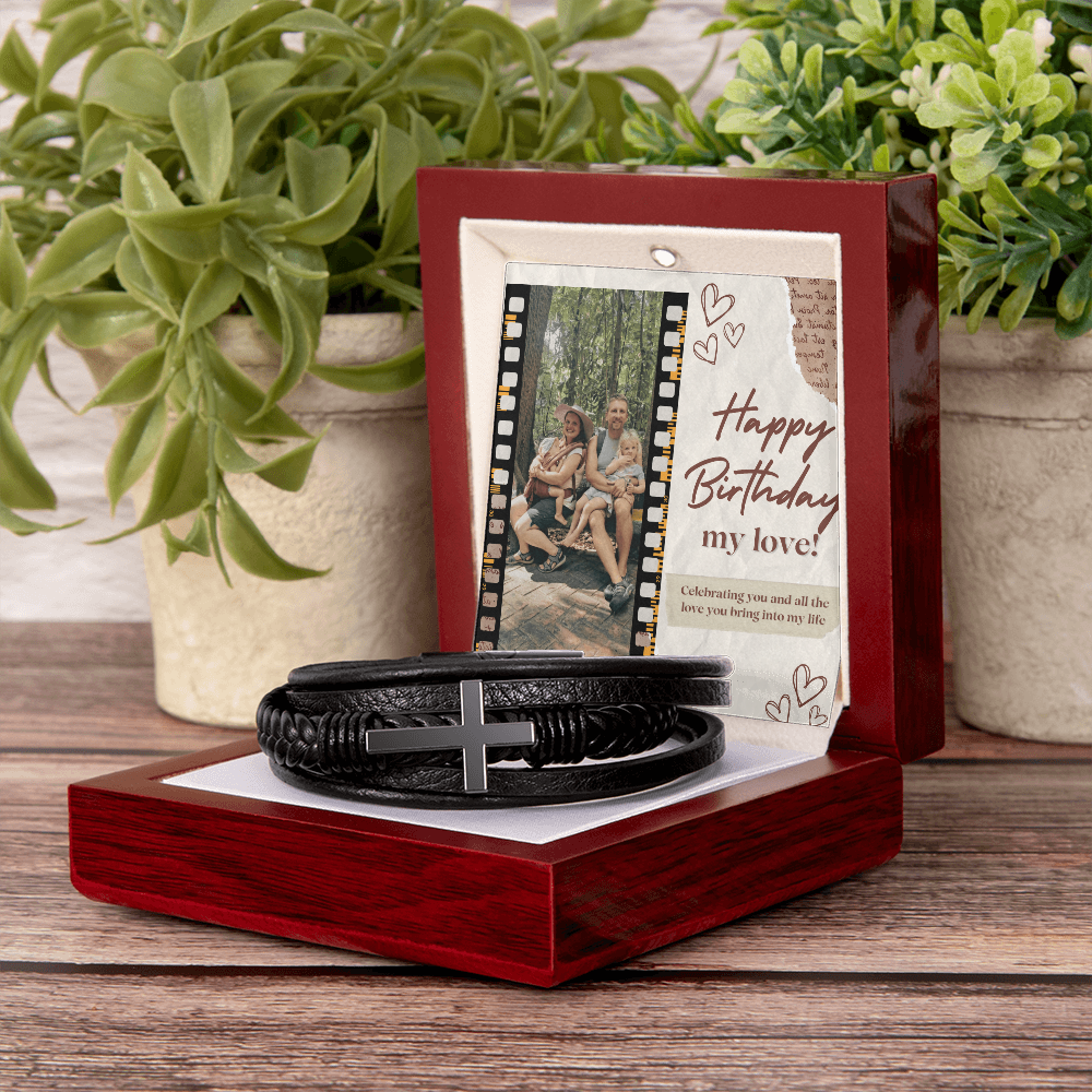 A black cross bracelet presented on a wooden box with a custom photo and message, displayed on a table with potted plants in the background.